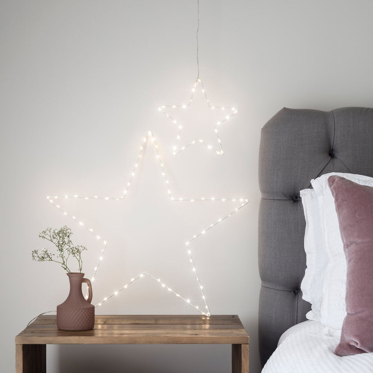 LED-Sterne leuchten neben einem Bett auf einem Holztisch mit brauner Vase und weißer Wand