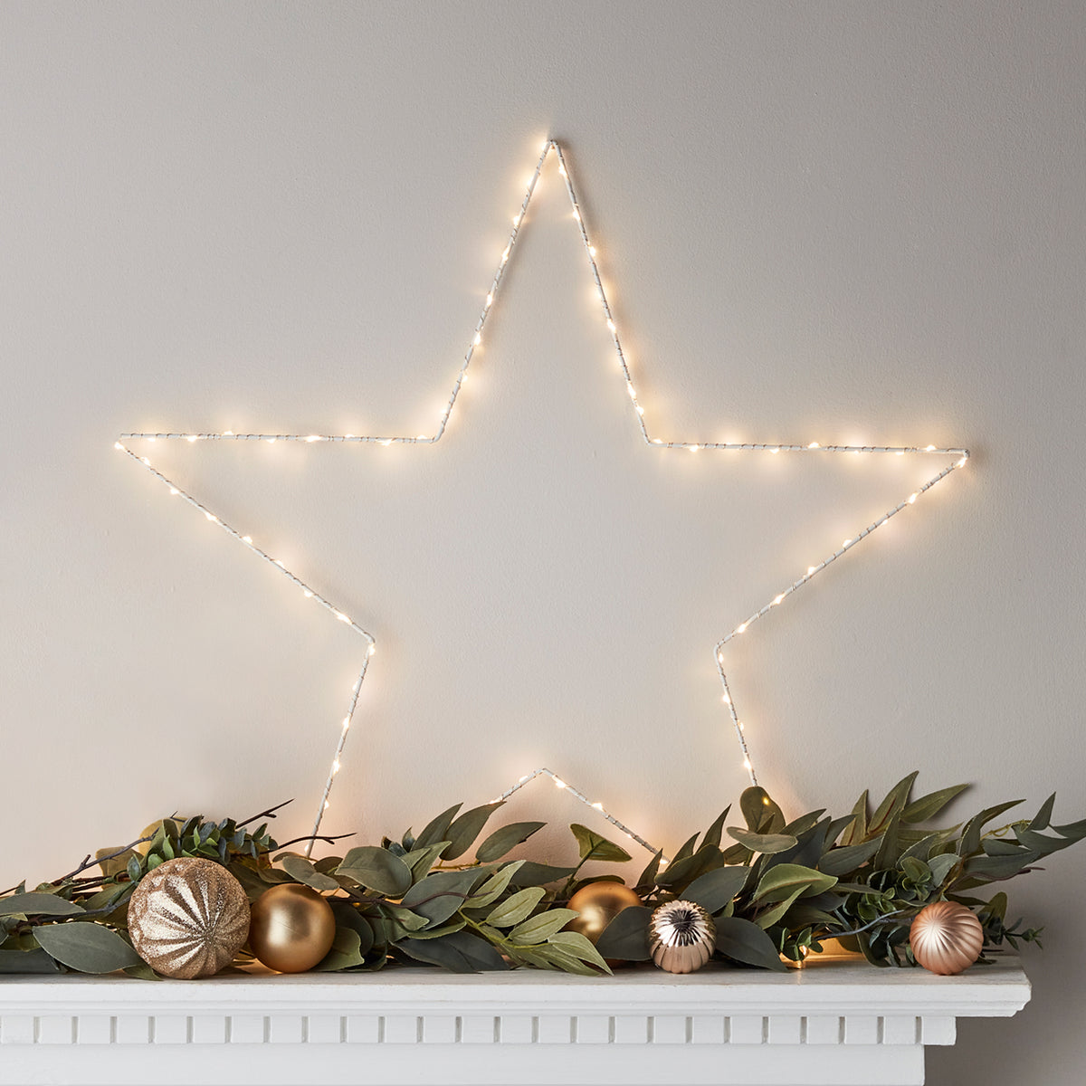 Leuchtender weißer LED-Stern mit warmweißen Micro LEDs an Wand über Dekoration mit grünen Blättern und goldenen Kugeln
