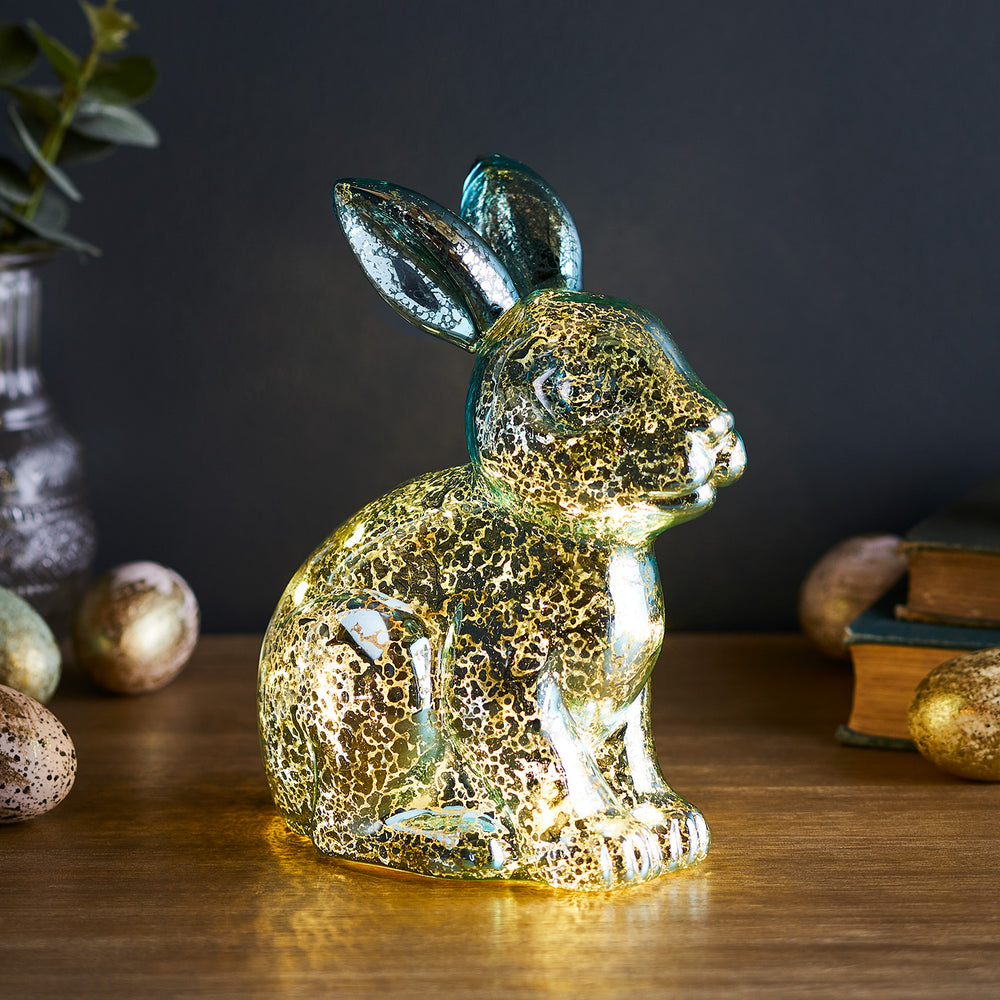 Beleuchteter Glas-Hase mit goldener Melierung auf Holztisch vor dunklem Hintergrund mit Osterdeko