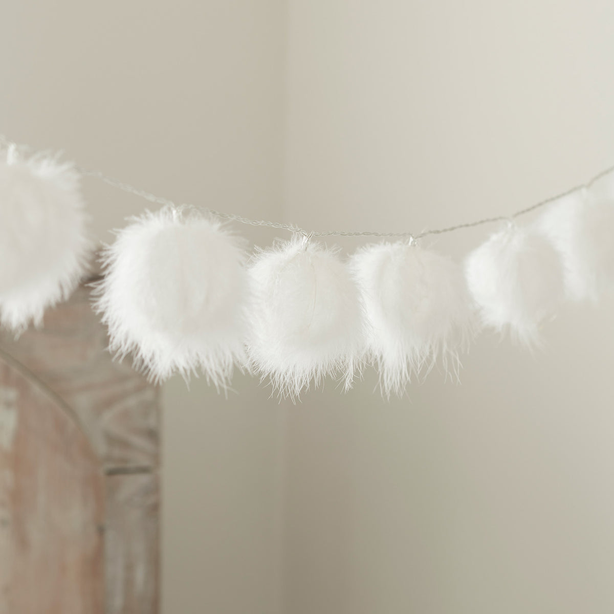 Weiße flauschige Bälle an einer Lichterkette, aufgehängt vor einer Wand mit Dekoration