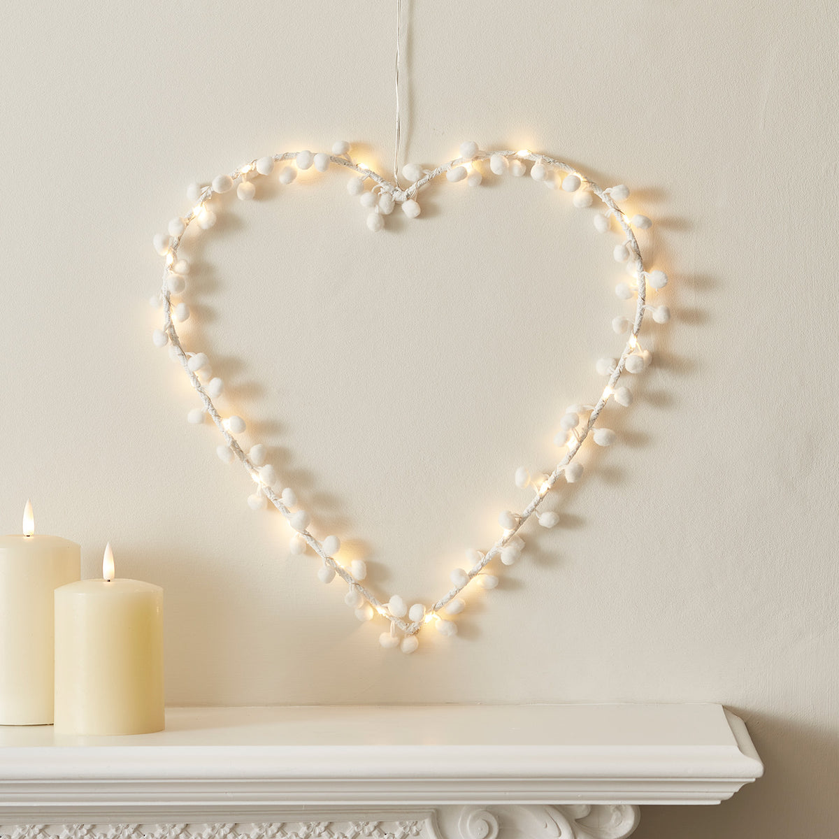 Weißes Micro LED Herz mit kleinen weißen Pompons an Wand über Kaminsims mit Kerzen