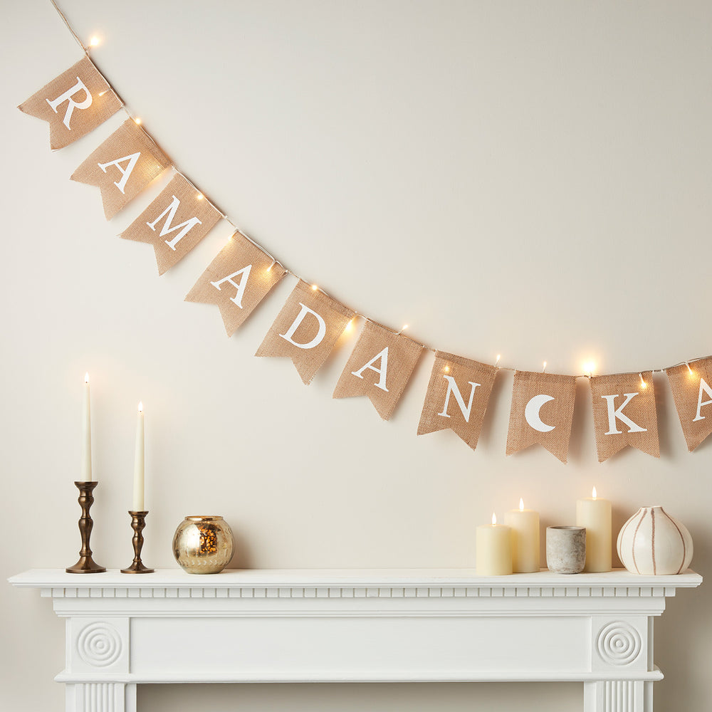 LED-Girlande mit Jutewimpeln und weißen Buchstaben RAMADAN an Wand über Kaminsims