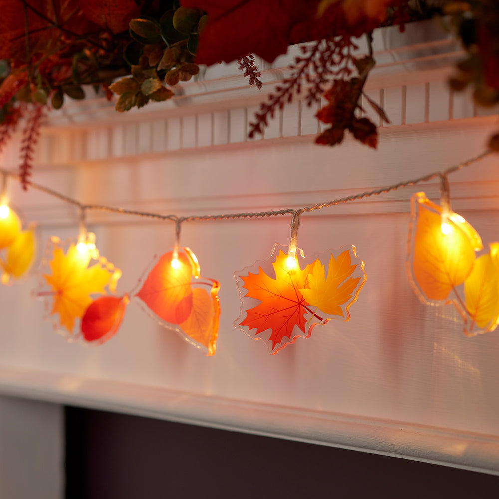 Lichterkette mit leuchtenden Acryl-Ahornblättern in warmem Gelb und Orange an weißem Kaminsims