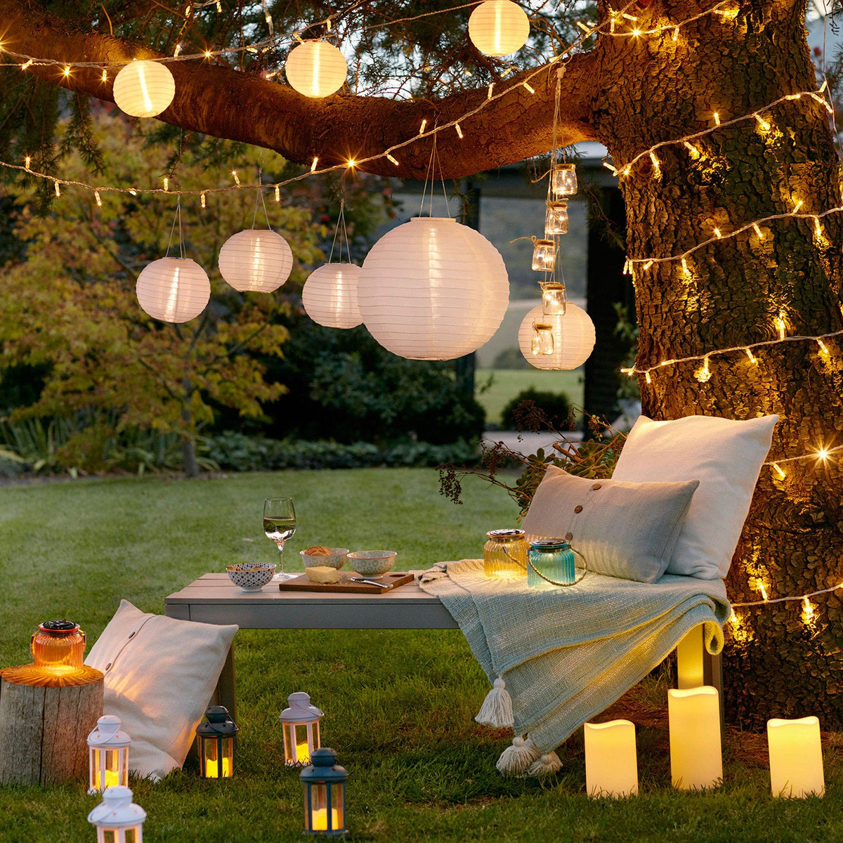 Garten mit hängenden Papierlaternen, LED-Lichterketten und Kerzen auf einer Bank