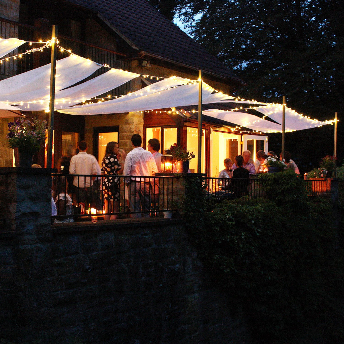 Abendliche Gartenparty mit LED-Lichterketten über weißem Sonnensegel und Gästen