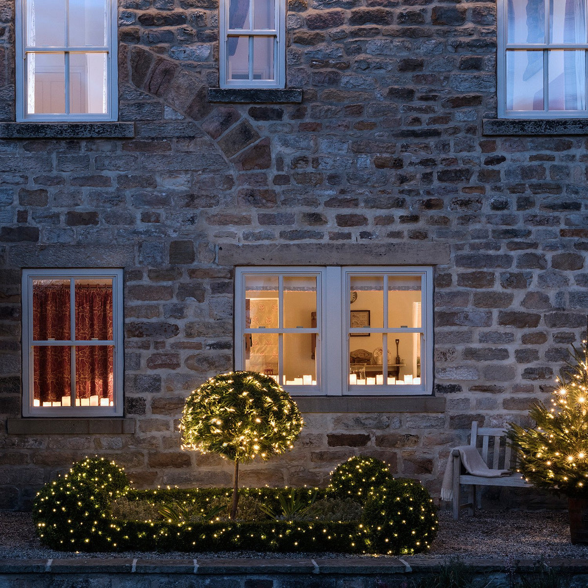 Beleuchtete Hecken und Kugelpflanzen mit warmweißen LEDs vor einem Haus mit Steinmauer
