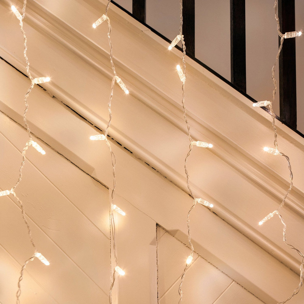 Warmweiße LED-Lichtervorhänge mit transparentem Kabel hängen vor einer weißen Wand und Treppe