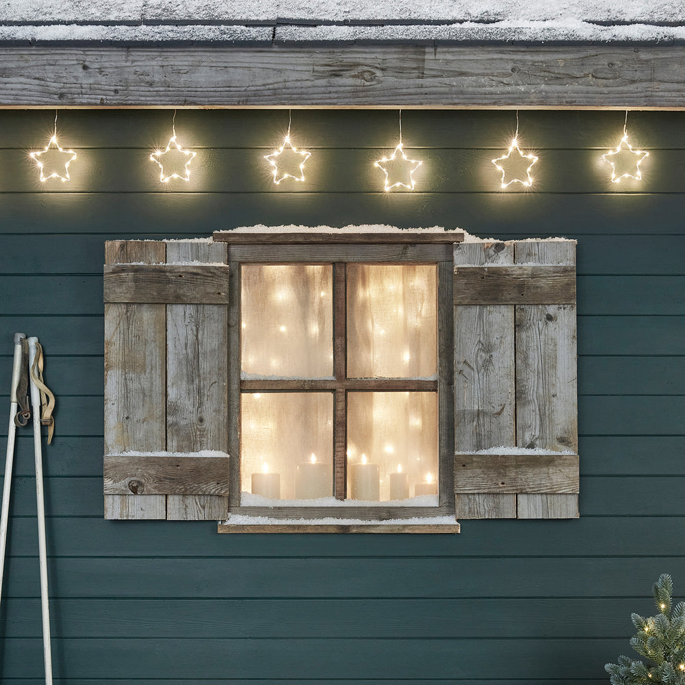 Sechs leuchtende LED-Sterne hängen über einem Fenster mit Holzläden und Schnee
