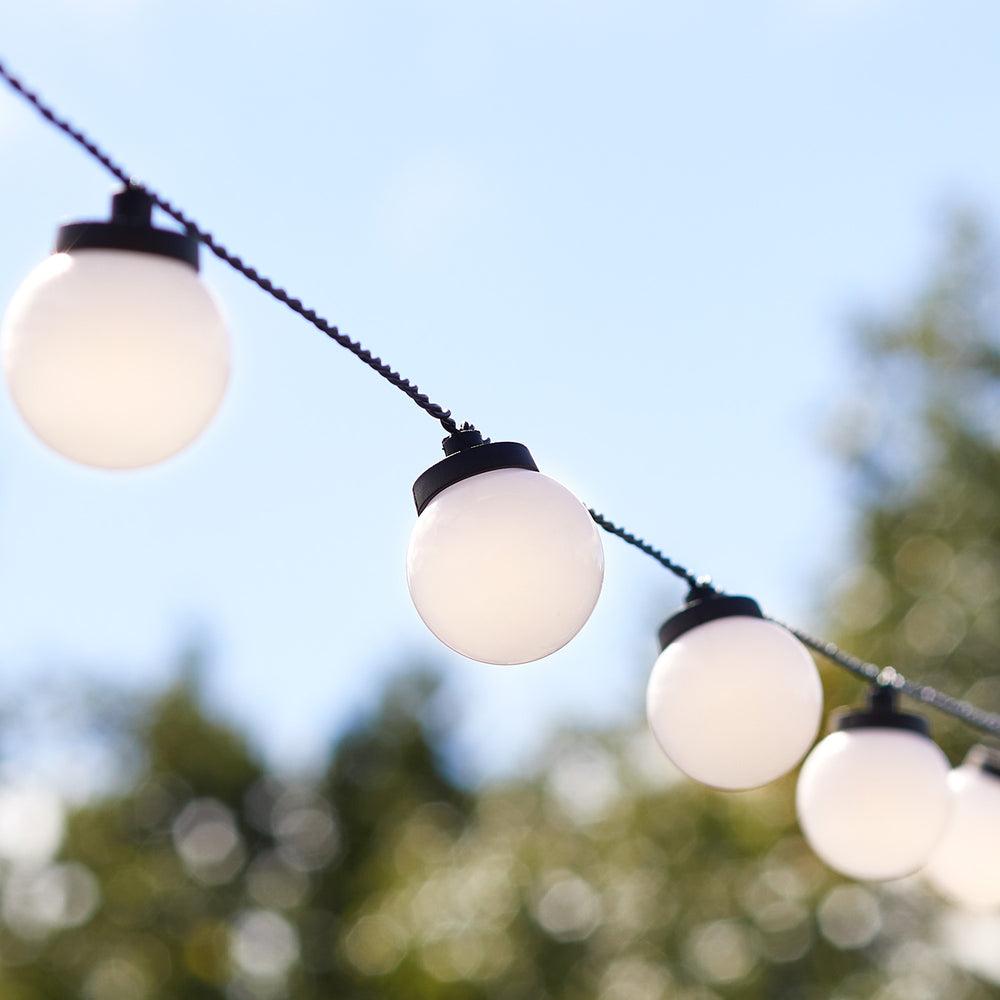 Nahaufnahme der weißen Kugellampen einer Lichterkette vor blauem Himmel und unscharfen Bäumen
