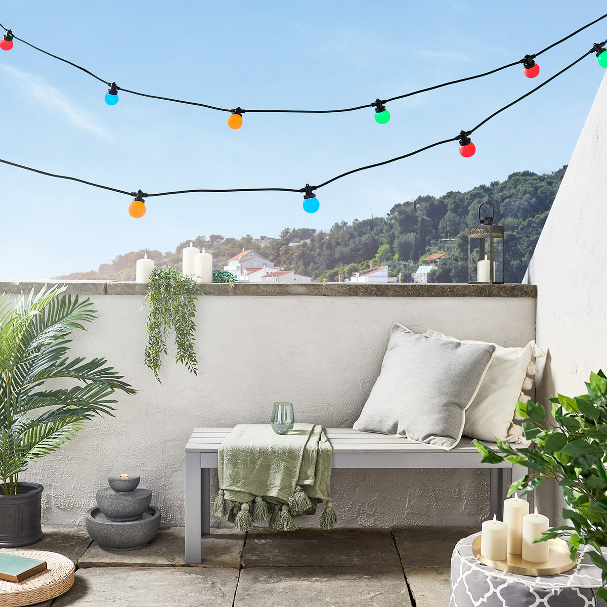 LED-Lichterkette bunt leuchtend mit rot, orange, blau und grün über grauer Bank auf Terrasse mit Kissen