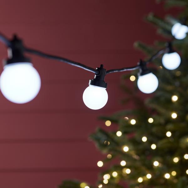 Weiße LED-Glühbirnen an schwarzem Kabel vor roter Wand und beleuchtetem Baum im Hintergrund