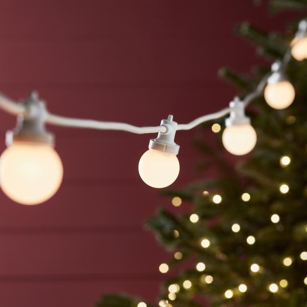 Warmweiße LED-Glühbirnen an weißem Kabel vor roter Wand und Weihnachtsbaum mit Lichtern