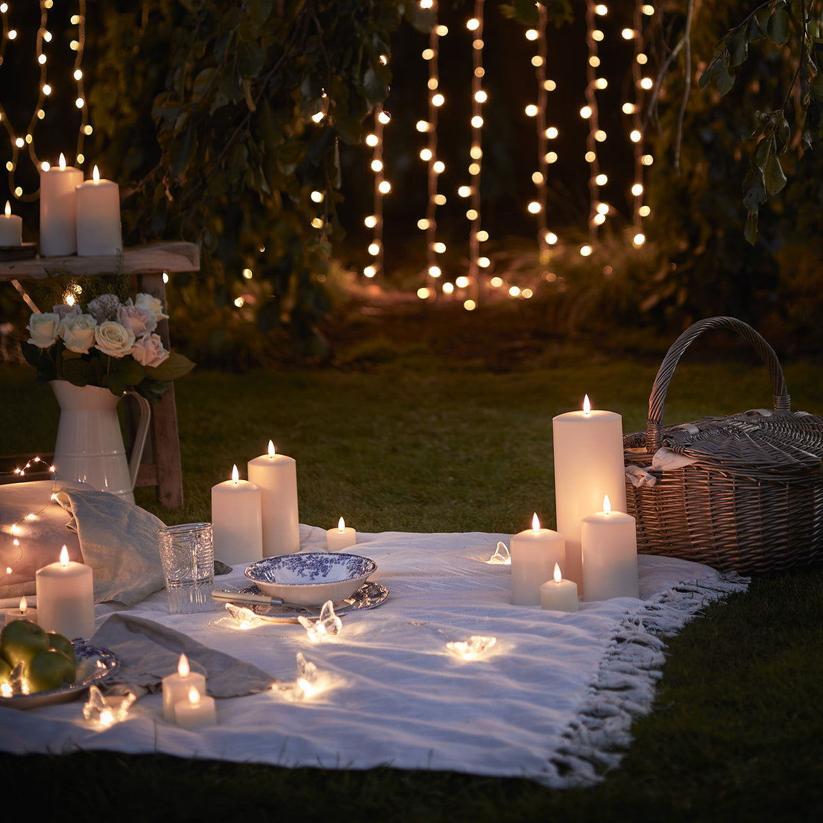 Mehrere LED-Kerzen und Lichterketten auf Picknickdecke im Garten bei Abenddämmerung