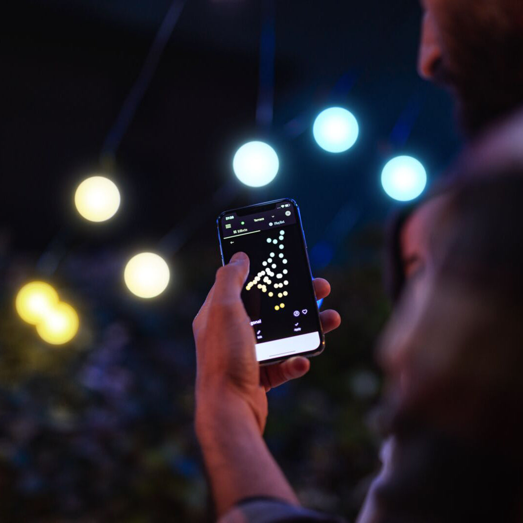 Person steuert smarte Party-Lichterkette mit Smartphone-App, LED-Glühbirnen leuchten im Hintergrund