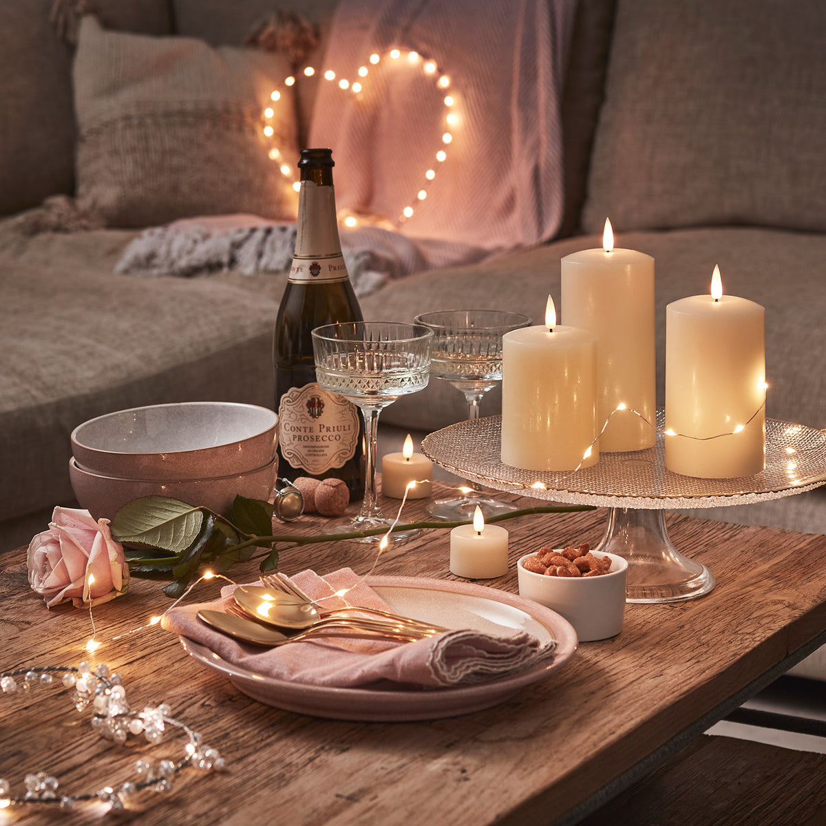 Gemütliche Tischdekoration mit Kerzen, Prosecco, Gläsern und roségoldener LED-Herzbeleuchtung im Hintergrund