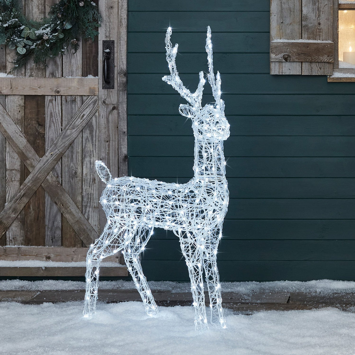 LED-Rentier mit weißen Lichtern vor grün gestrichener Holzfassade und Schnee