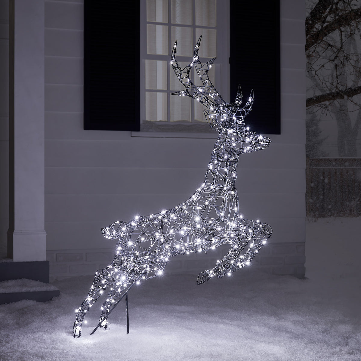 Springendes LED-Rentier mit weißen Lichtern vor Hausfassade im Schnee